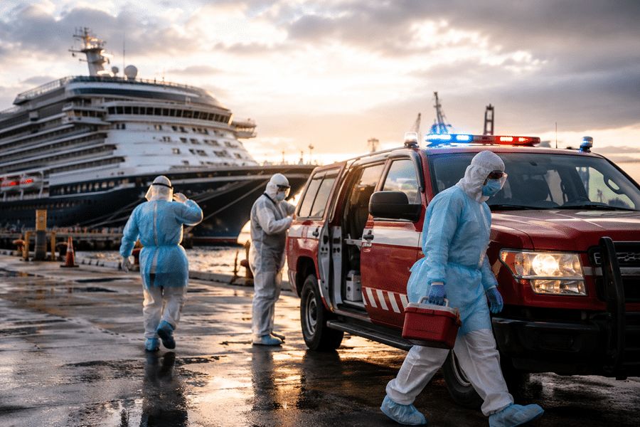 Hazmat-suited medical team unloads at a dock beside a large cruise ship and a red emergency vehicle with flashing lights, sunset backdrop.